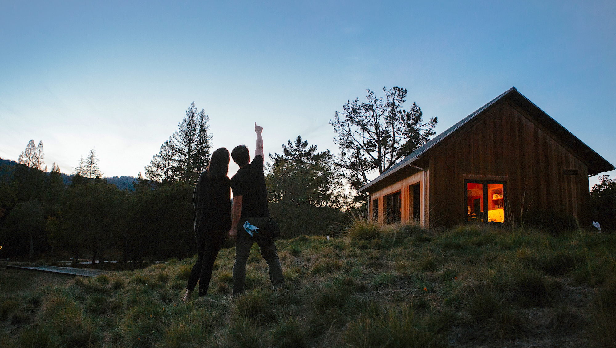 Two people outside of a log cabin, facing away from the camera and pointing towards the sky at dusk